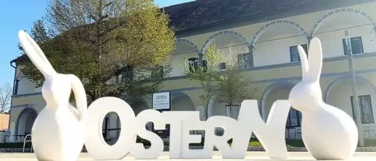 Ein großes weißes Schild, das Ostern buchstabiert, steht vor einem Gebäude mit Bogenfenstern. Im Hintergrund sind Bäume und ein klarer Himmel zu sehen.