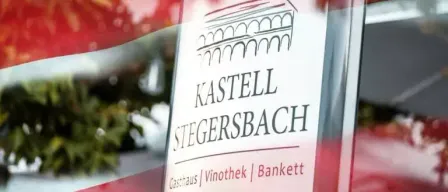 Ein Schild mit 'Kastell Stegersbach' und 'Gasthaus, Vinothek, Bankett' wird auf einer transparenten Oberfläche angezeigt. Das Schild ist von Rot umrahmt, was möglicherweise auf eine Veranstaltung oder einen besonderen Anlass hinweist.