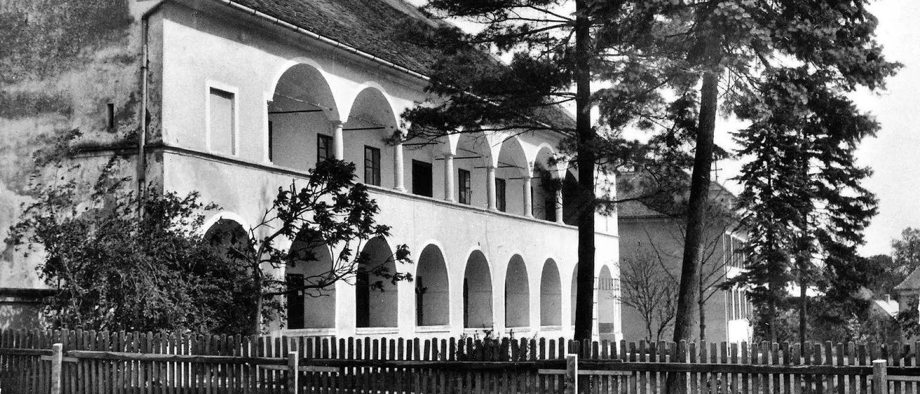 Ein Schwarz-Weiß-Bild eines alten Gebäudes mit Bogenfenstern und einem gefliesten Dach. Vor dem Gebäude befinden sich Bäume und ein Holzzaun.