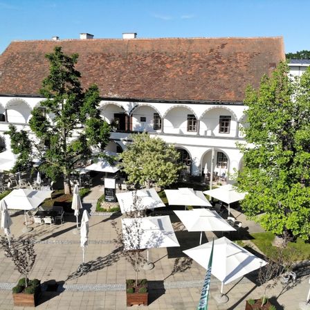 Eine Vogelperspektive auf ein Gebäude mit braunem Dach und weißen Wänden. Der Innenhof ist mit weißen Sonnenschirmen und Stühlen gefüllt, und es gibt Bäume und Pflanzen in der Umgebung.