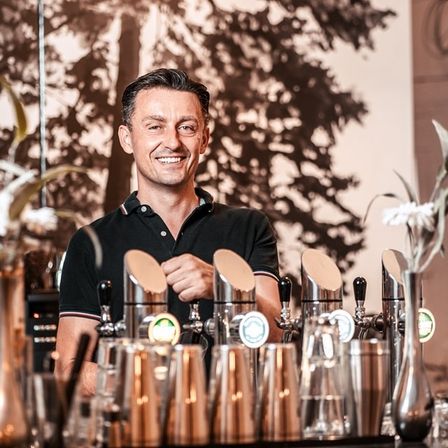 Ein lächelnder Mann steht hinter einer Bar mit verschiedenen Cocktail-Shakern und Gläsern. Er trägt ein schwarzes Poloshirt. Hinter ihm befindet sich ein verschwommenes Bild eines Baumes und einer Wand.