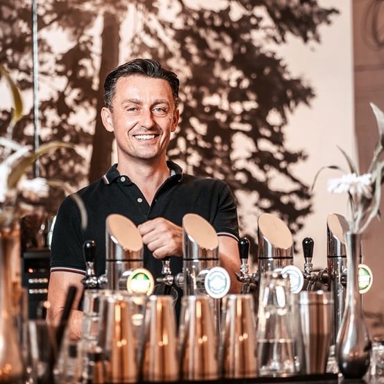 Ein lächelnder Mann steht hinter einer Bar mit verschiedenen Cocktail-Shakern und Gläsern. Er trägt ein schwarzes Poloshirt. Hinter ihm befindet sich ein verschwommenes Bild eines Baumes und einer Wand.