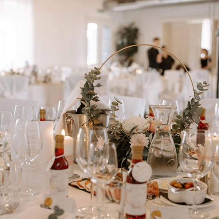 Ein formeller Esstisch mit Weingläsern, Kerzen, Flaschen und einem Korb mit Grünpflanzen. Der Tisch ist mit einem weißen Tuch bedeckt. Im Hintergrund stehen Leute.