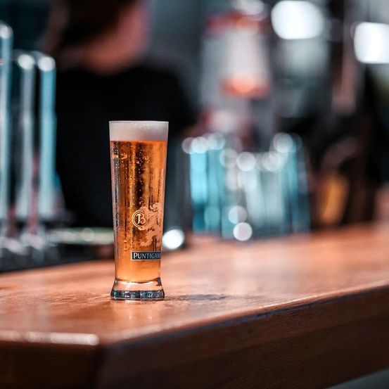 Ein Glas Puntigam-Bier steht auf einer hölzernen Theke, mit einem verschwommenen Hintergrund eines Barbereichs.