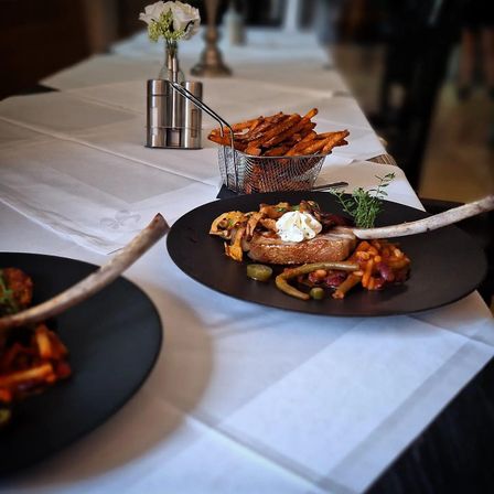 Zwei schwarze Teller mit einer Mahlzeit und einem Korb mit Pommes Frites auf einem Tisch mit weißer Tischdecke. Ein Vasen mit weißen Blumen ist auf dem Tisch platziert.