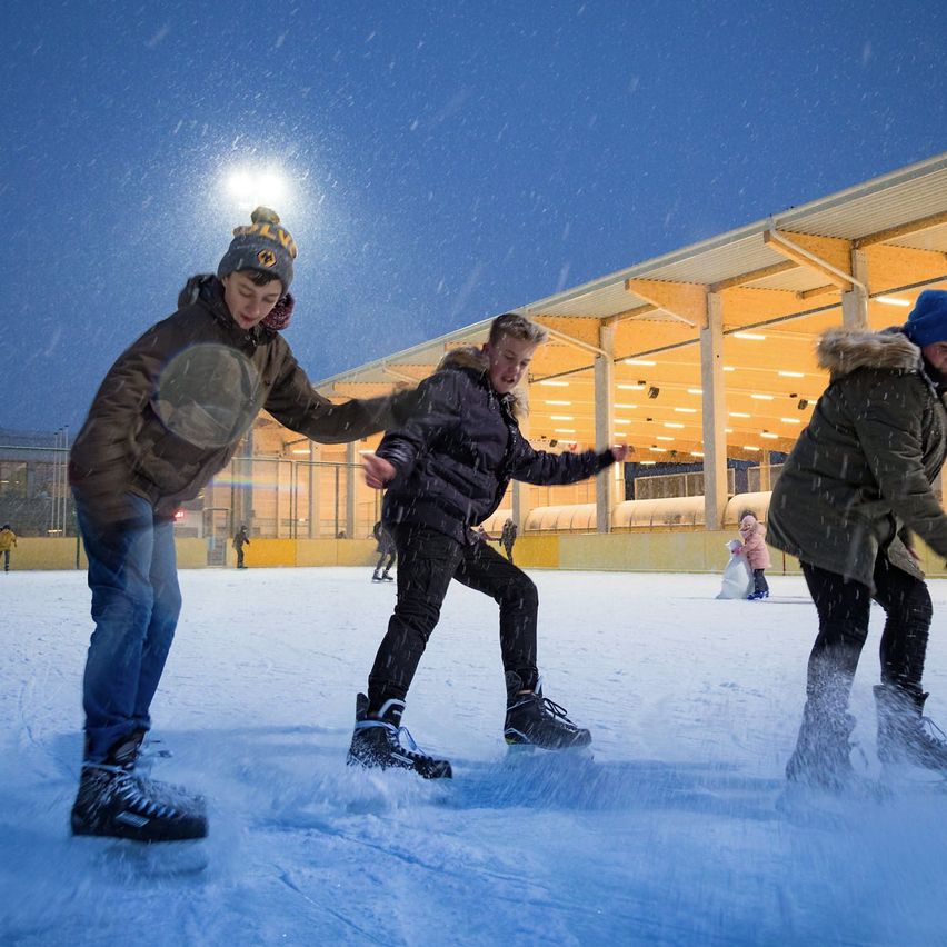 Drei Jungen skaten auf einer Eisbahn im Schnee. Ein Gebäude mit gelben Lichtern steht dahinter.