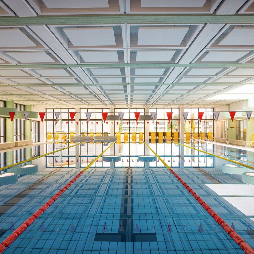 Ein leerer Hallenschwimmbad mit blauen Kacheln, roten Bahnen und überhängenden Flaggen. Der Pool ist gut beleuchtet, und die Fenster reflektieren den Pool.
