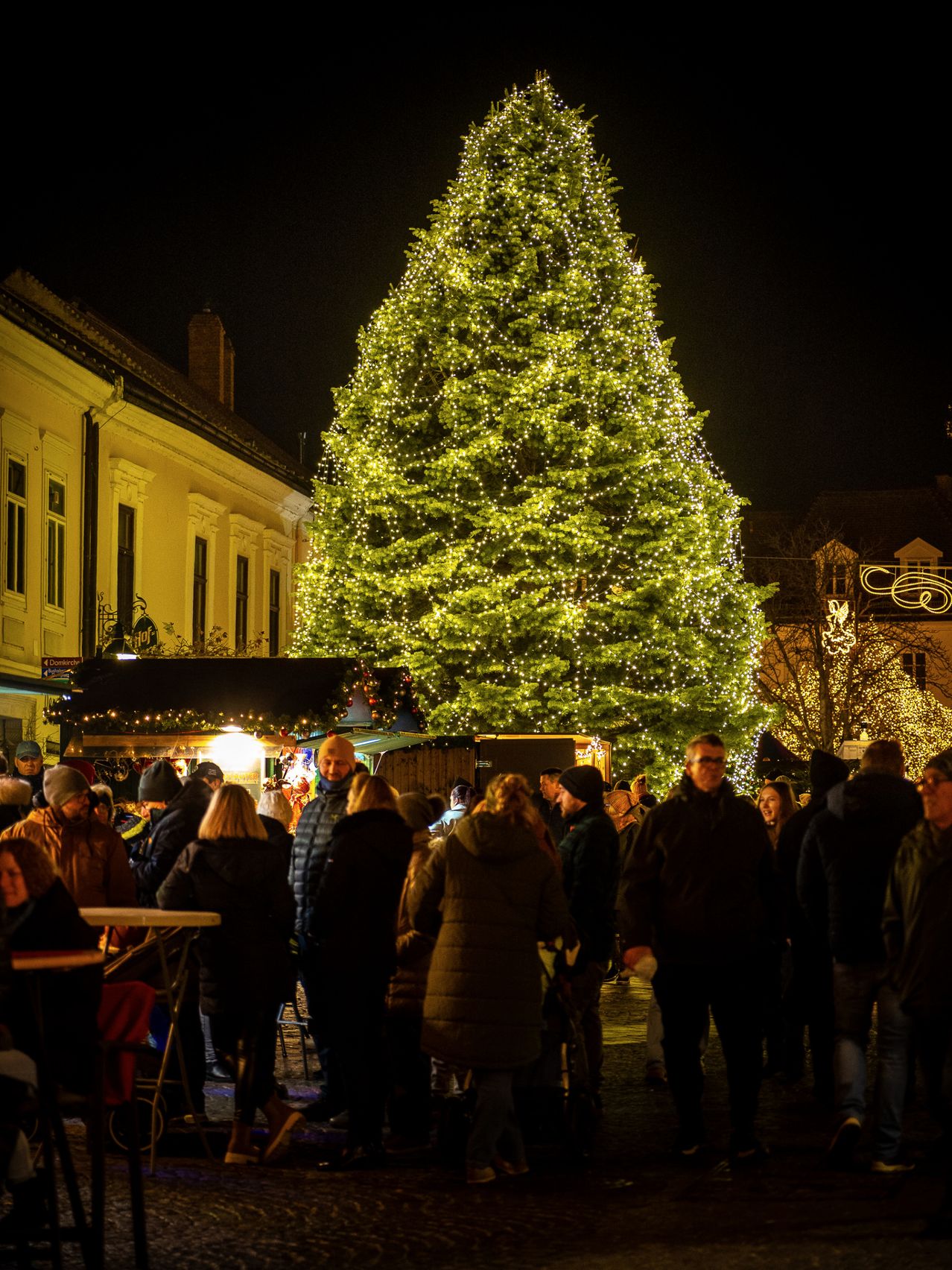Ein großer Weihnachtsbaum ist beleuchtet und von Menschen umgeben, wahrscheinlich auf einem Weihnachtsmarkt. Im Hintergrund befinden sich Gebäude mit mehreren Fenstern und dekorativen Lichtern.