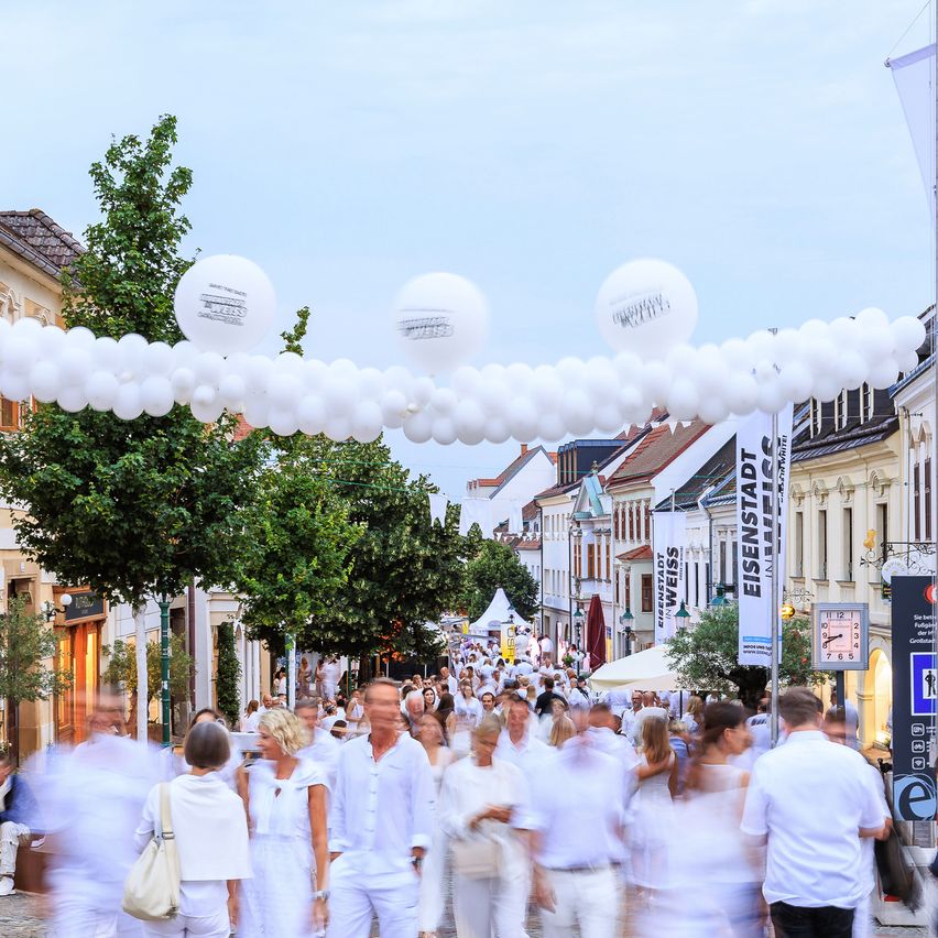 Eine belebte Straße voller Menschen in Weiß, unter einem dekorativen Bogen aus weißen Luftballons, mit Geschäften und Bäumen im Hintergrund.