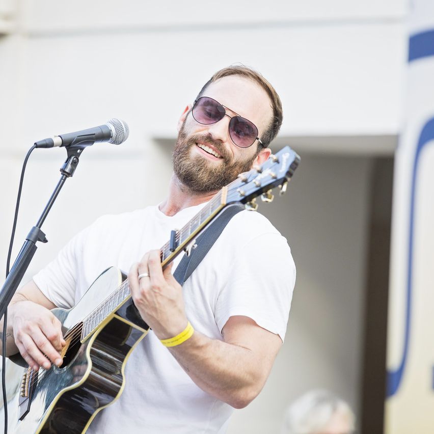 Ein bärtiger Mann in einem weißen T-Shirt und Sonnenbrille spielt Gitarre und singt in ein Mikrofon.