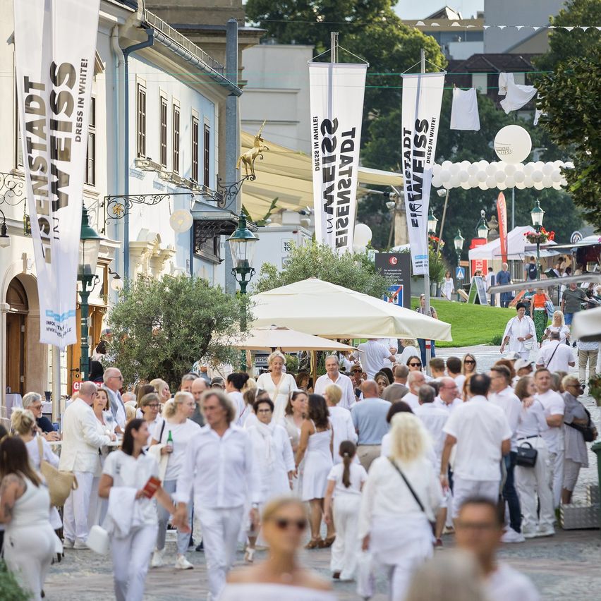 Eine belebte Straße mit Menschen in Weiß, Straßenbannern und Straßenlampen. Im Hintergrund befinden sich Schirme, Bäume und Fahnen.