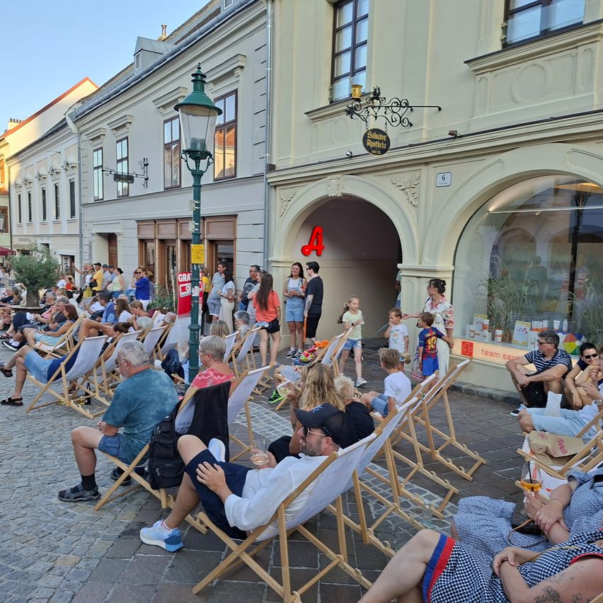 Eine Menschenmenge sitzt auf Klappstühlen auf einer Kopfsteinpflasterstraße, einige schauen einen Film, andere plaudern. Gebäude mit Glasfenstern und eine Straßenlaterne sind im Hintergrund.
