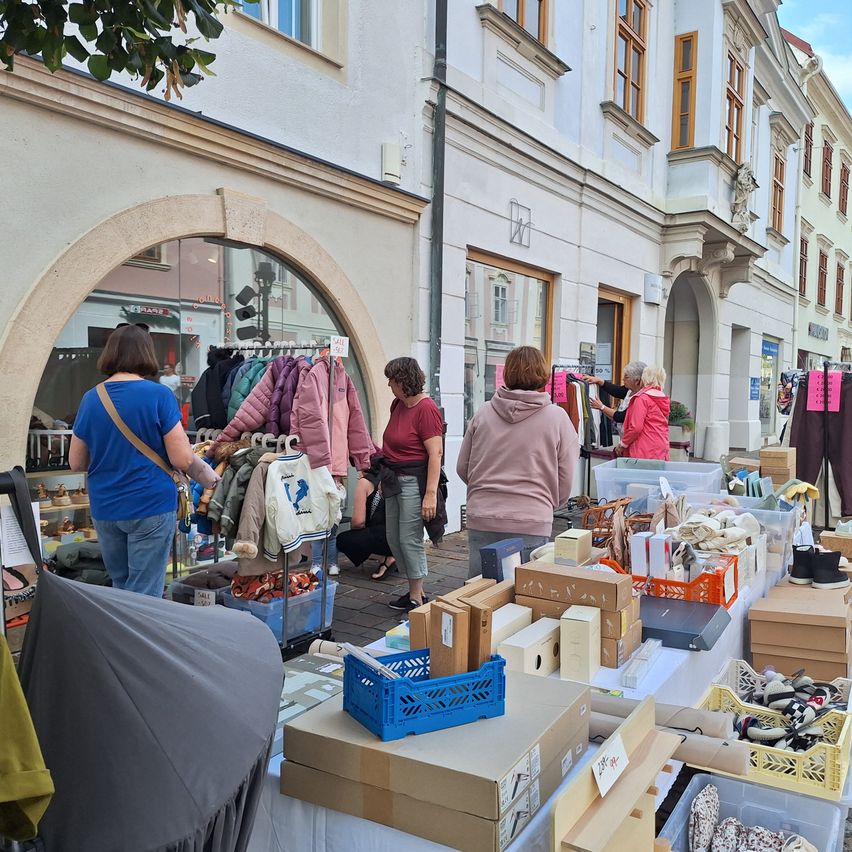 A street market with various items on display, including clothing and accessories, in front of a building with multiple windows. People are browsing the items.