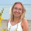 Eine Frau mit blonden Haaren und Brille posiert für ein Foto am Strand, umgeben von gelben Blumen.