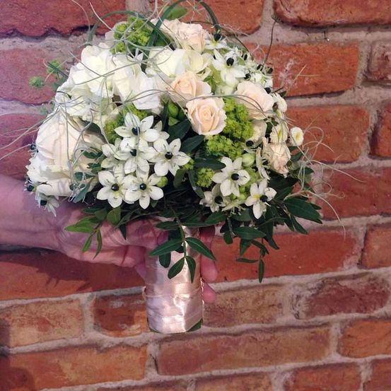 Ein Blumenstrauß mit weißen Blüten und grünen Blättern, gebunden mit einem rosa Band, vor einer Ziegelwand gehalten.