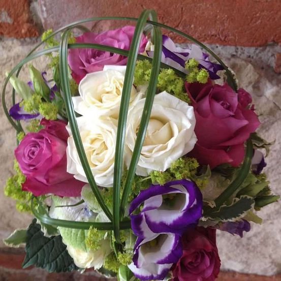 Ein kugelförmiges Blumenarrangement mit weißen, rosa und lila Rosen, ergänzt durch grüne Blätter und Blumen, lehnt sich gegen eine Ziegelwand.