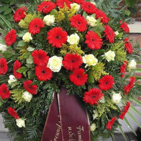 Ein Blumenarrangement mit roten und weißen Blumen und Grün, mit einer braunroten Schleife mit weißem Text, möglicherweise für eine Gedenkfeier.