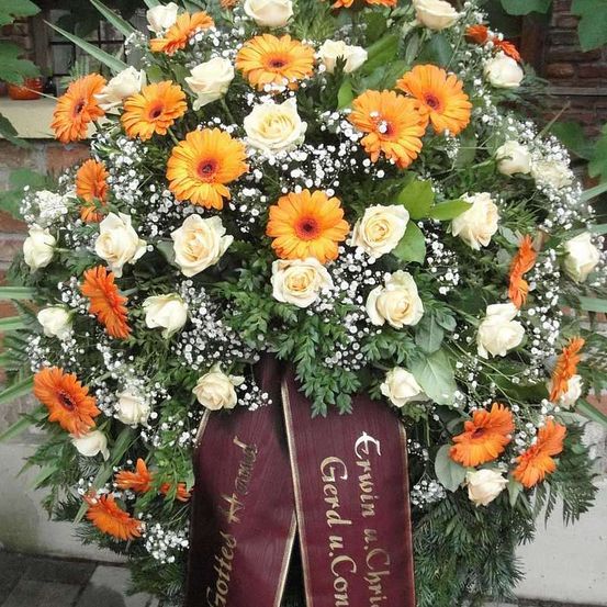 Eine Blumengestecke aus weißen und orangefarbenen Blumen mit grünen Blättern und einem Band, das Erwin u. Christa Gerd u. Conni Gottle Afrand liest.