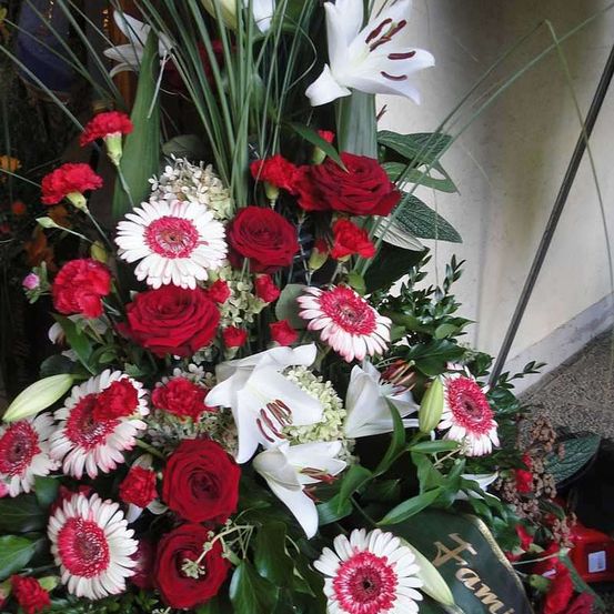 Eine große Blumenarrangement besteht aus roten und weißen Blumen, einschließlich Rosen und Gänseblümchen, mit grünen Blättern und einer weißen Lilie. Das Arrangement wird an einer hellen Wand ausgestellt.