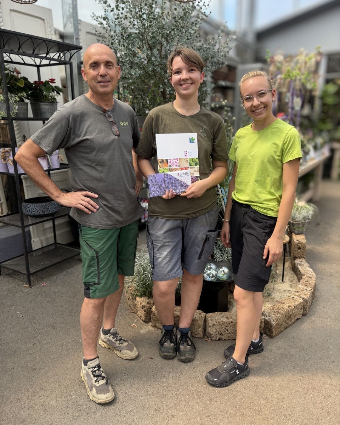 Drei Personen stehen in einem Gartencenter, lächeln und halten ein Buch. Der Mann auf der linken Seite trägt ein graues T-Shirt und grüne Shorts. Die mittlere Person, eine Frau, hält ein Buch mit buntem Cover. Die Frau auf der rechten Seite trägt ein grünes T-Shirt und schwarze Shorts.