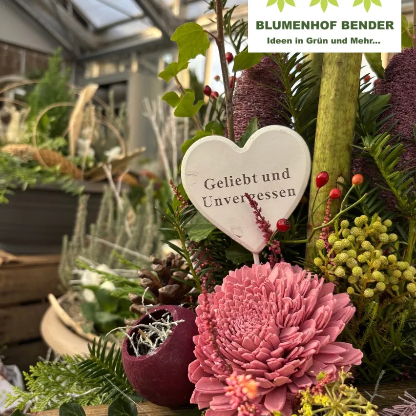 Eine Blumenarrangement in einem Gewächshaus mit einem herzförmigen Schild, das 'Geliebt und Unvergessen' besagt.
