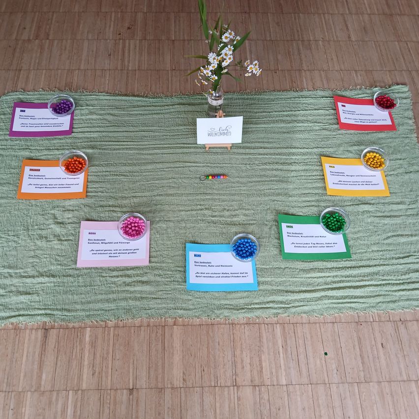 Ein grüner Stoff mit verschiedenen farbigen Perlen und kleinen Karten mit Text, eine Blume in einer Vase und ein Schild, das 'Liebe Willkommen' sagt.
