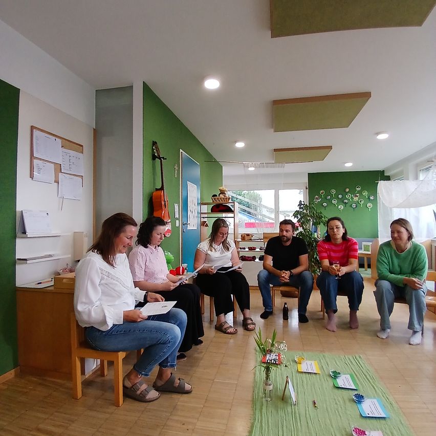 Eine Gruppe von Menschen sitzt im Kreis in einem Raum mit grünen Wänden, liest Papiere und diskutiert. Eine Gitarre hängt an der Wand und eine Pflanze steht auf dem Tisch.
