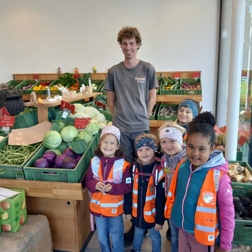 Ein Mann steht hinter einer Gruppe von Kindern in orangefarbenen Westen und lächelt vor einem Gemüsestand.