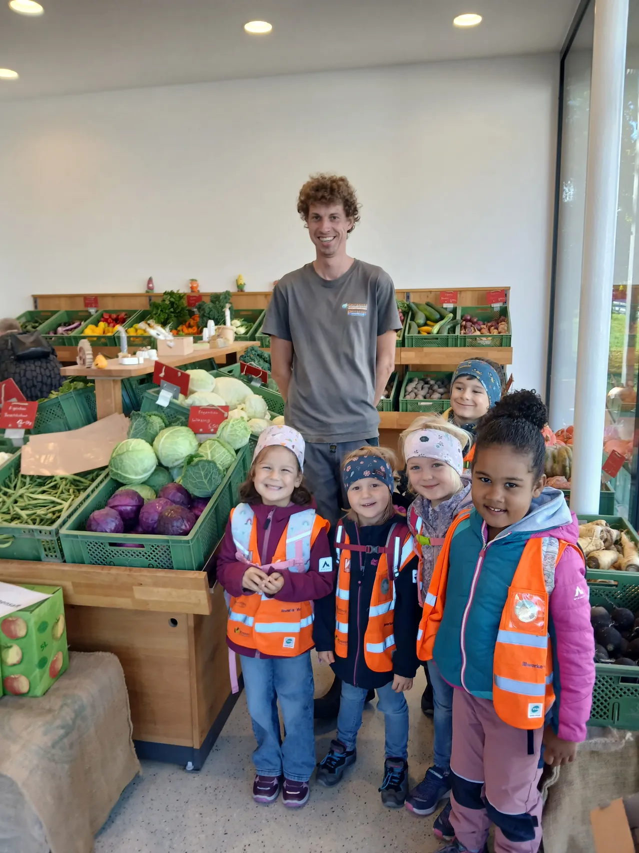 Ein Mann steht hinter einer Gruppe von Kindern in orangefarbenen Westen und lächelt vor einem Gemüsestand.