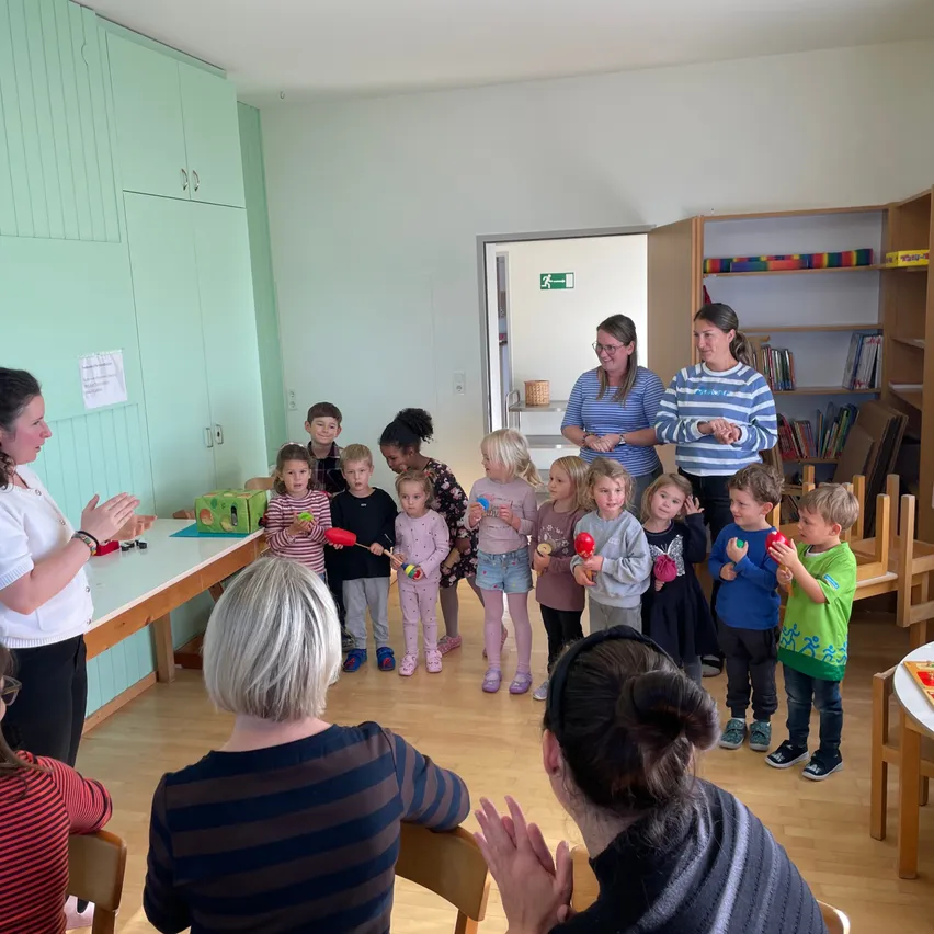 Eine Gruppe von Kindern und Erwachsenen versammelt sich in einem Raum, möglicherweise für eine musikalische Aktivität. Die Kinder halten rote Rasseln und scheinen der Frau vor ihnen zuzuhören.