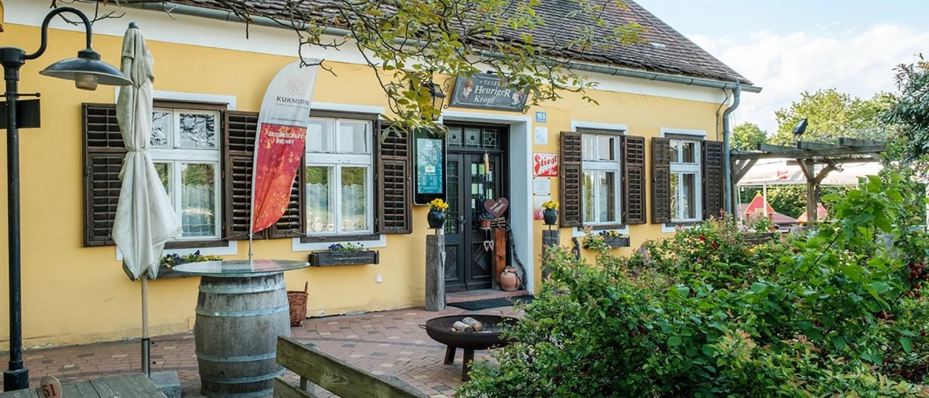 Ein gelbes Gebäude mit braunen Fensterläden hat ein Banner. Es hat eine schwarze Tür mit einem Schild darüber. Es gibt Pflanzen, einen Fass und einen Feuerkorb.