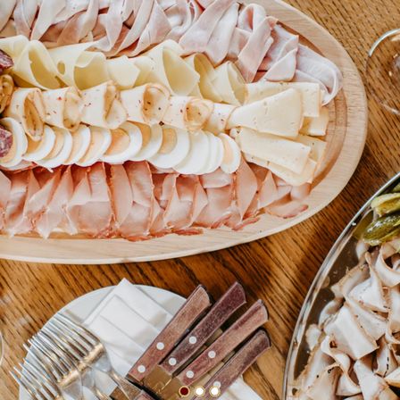 Ein Holztisch zeigt ein großes Tablett mit verschiedenen Fleisch- und Käsesorten, begleitet von Gabeln und Messern auf einem weißen Teller. Eine weitere Schale enthält eingelegtes Gemüse.