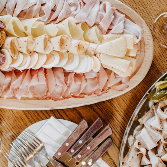 Ein Holztisch zeigt ein großes Tablett mit verschiedenen Fleisch- und Käsesorten, begleitet von Gabeln und Messern auf einem weißen Teller. Eine weitere Schale enthält eingelegtes Gemüse.