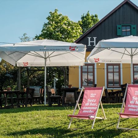 Ein Restaurant mit Außenbestuhlung unter großen weißen Sonnenschirmen. Ein Rasenbereich mit Klappstühlen, auf denen 'Sitz di her, samma mehr' steht. Bäume und Pflanzen im Hintergrund.