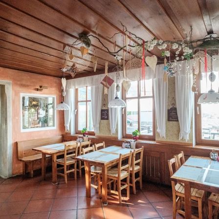 Ein leeres Holzrestaurant mit mehreren Tischen und Stühlen, geschmückt mit Dekorationen, weißen Vorhängen und Topfpflanzen. Die Decke hat hängende Dekorationen und Lampen.