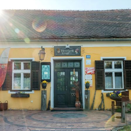 Ein hellgelbes Gebäude mit schwarzen Fensterläden, einer Tür mit Herzdesign und Topfpflanzen. Der Name 'Heuriger Kropf' steht über der Tür. Eine Sonne scheint von links.