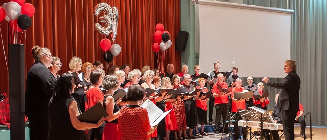 Ein Chor tritt auf der Bühne auf, mit einer Projektionsleinwand im Hintergrund und Luftballons. Die Chormitglieder sind in Rot gekleidet, während der Dirigent davor steht.