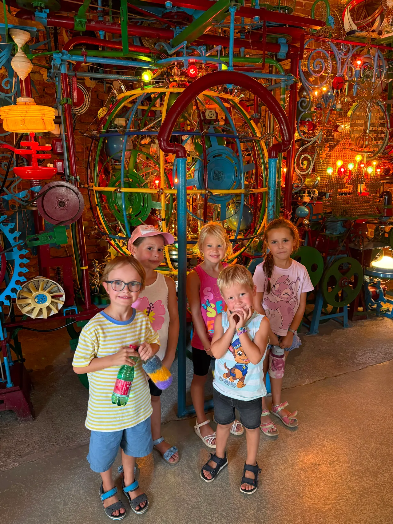 Fünf Kinder stehen vor einer farbenfrohen, komplizierten mechanischen Installation und halten verschiedene Gegenstände in den Händen. Im Hintergrund befindet sich ein farbenfrohes Labyrinth aus Rohren und Zahnrädern.