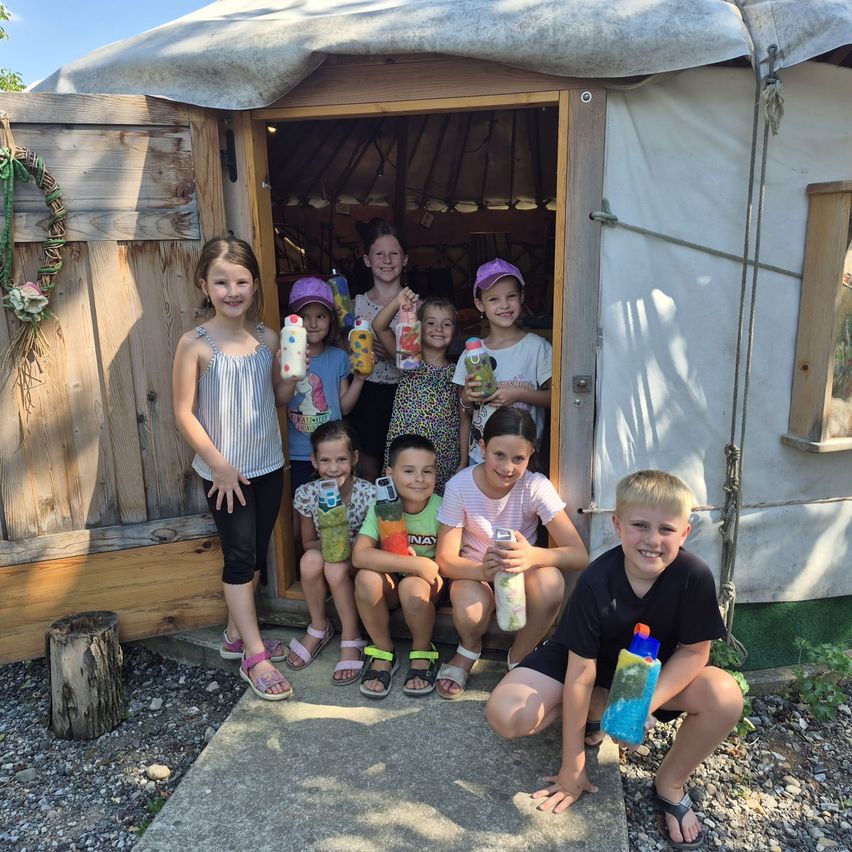 Eine Gruppe von Kindern lächelt und posiert für ein Foto vor einem Jurte, jeder hält eine Flasche. Die Jurte hat Holzwände und eine Holztür.