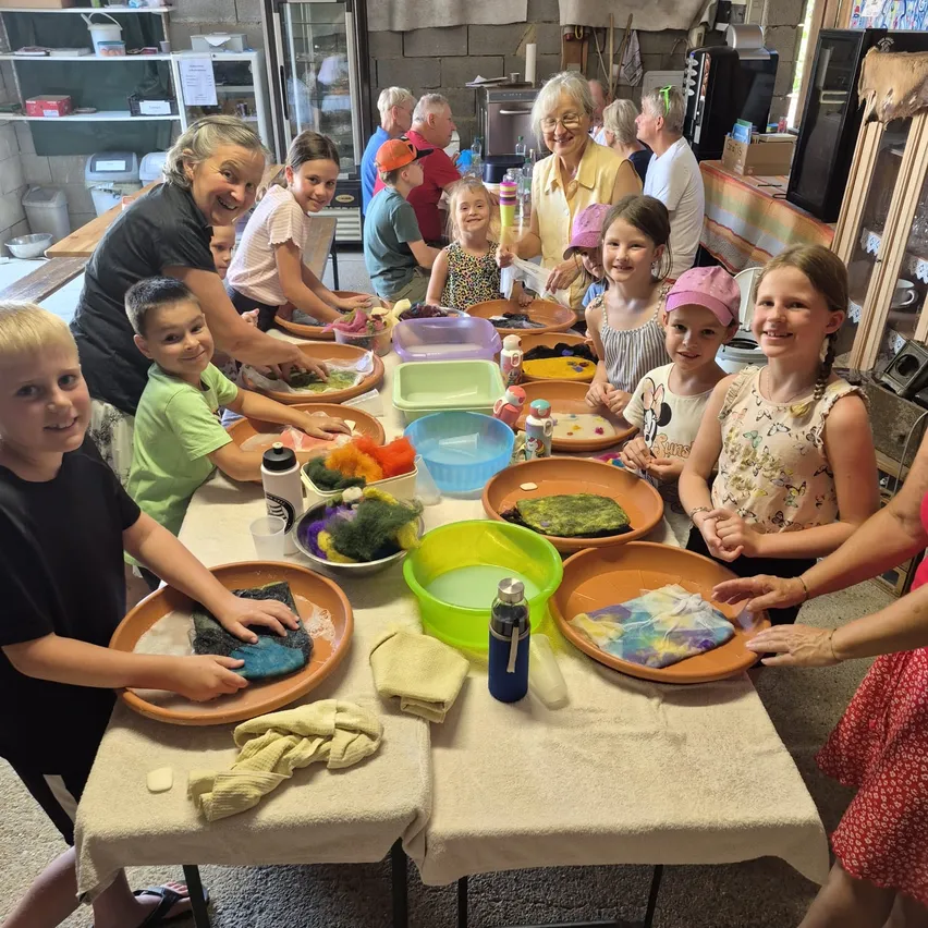 Eine Gruppe von Kindern und Erwachsenen ist um einen Tisch versammelt und beschäftigt sich mit einer Bastelaktivität. Sie arbeiten mit Materialien in Schüsseln und einige tragen Mützen. Der Tisch ist mit einem weißen Tuch bedeckt und hat verschiedene Gegenstände darauf.