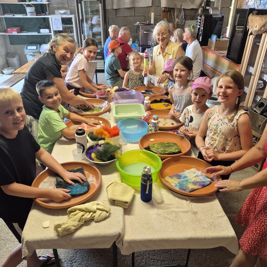 Eine Gruppe von Kindern und Erwachsenen ist um einen Tisch versammelt und beschäftigt sich mit einer Bastelaktivität. Sie arbeiten mit Materialien in Schüsseln und einige tragen Mützen. Der Tisch ist mit einem weißen Tuch bedeckt und hat verschiedene Gegenstände darauf.