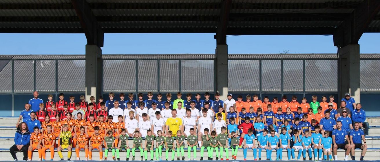 Eine Gruppe junger Fußballspieler in verschiedenen Trikotfarben posiert für ein Teamfoto in einem Stadion.