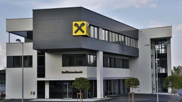 Die Vorderseite eines modernen Bankgebäudes mit einem gelben Kreuzlogo auf einer schwarzen Tafel, mit Glasfenstern und einem Schild, das 'Raiffeisenbank' steht. Zwei kleine Bäume stehen davor und Straßenlaternen sind auf beiden Seiten.