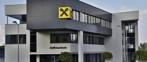 Die Vorderseite eines modernen Bankgebäudes mit einem gelben Kreuzlogo auf einer schwarzen Tafel, mit Glasfenstern und einem Schild, das 'Raiffeisenbank' steht. Zwei kleine Bäume stehen davor und Straßenlaternen sind auf beiden Seiten.