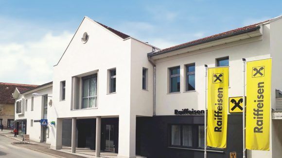 Ein weißes Gebäude mit rotem Dach, mehreren Fenstern und drei gelben Bannern mit der Aufschrift 'Raiffeisen' steht an einer Straße. Es gibt weitere Gebäude und eine Person, die in der Nähe geht.