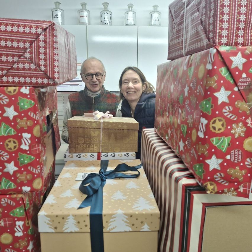 Zwei Personen stehen hinter einem Tisch mit mehreren Weihnachtsgeschenken und lächeln für ein Foto. Der Mann trägt eine Brille und einen Pullover, die Frau eine Jacke. Dahinter stehen weitere Weihnachtsgeschenke auf Regalen.