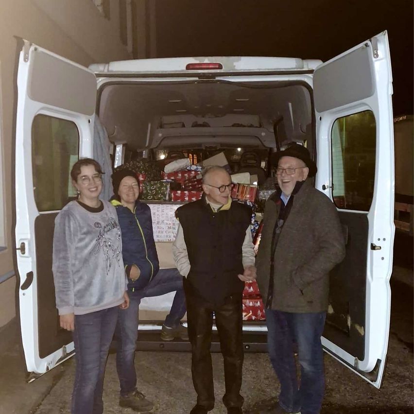 Vier Personen stehen vor einem weißen Lieferwagen mit offenen Türen. Sie scheinen Geschenke zu verteilen, da mehrere verpackte Geschenke im Inneren des Fahrzeugs zu sehen sind.