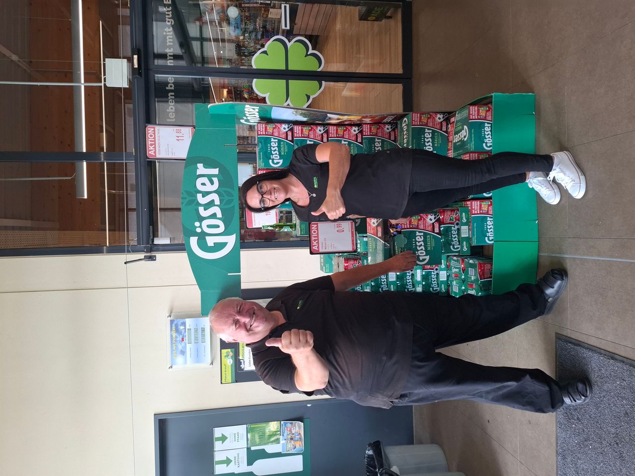Two employees of a store are posing for a photo in front of a display of Güsser beer. The woman is standing on a stack of beer boxes, and the man is giving a thumbs up.