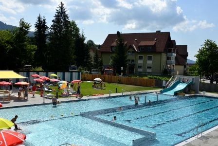 Ein Freibad mit Schwimmern und einem Gebäude im Hintergrund. Bunte Schirme und eine Rutsche sind um den Pool herum aufgebaut.