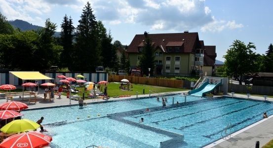 Ein Freibad mit Schwimmern und einem Gebäude im Hintergrund. Bunte Schirme und eine Rutsche sind um den Pool herum aufgebaut.